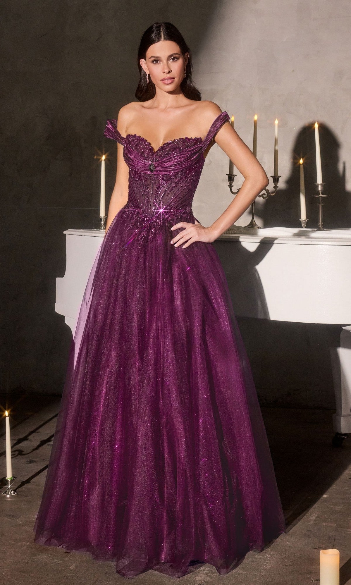 A woman in the Ladivine CC493 Long Prom Dress in eggplant, featuring an off-shoulder sweetheart neckline, stands by a white piano with lit candles, her elegant silhouette highlighted by the dimly lit room.